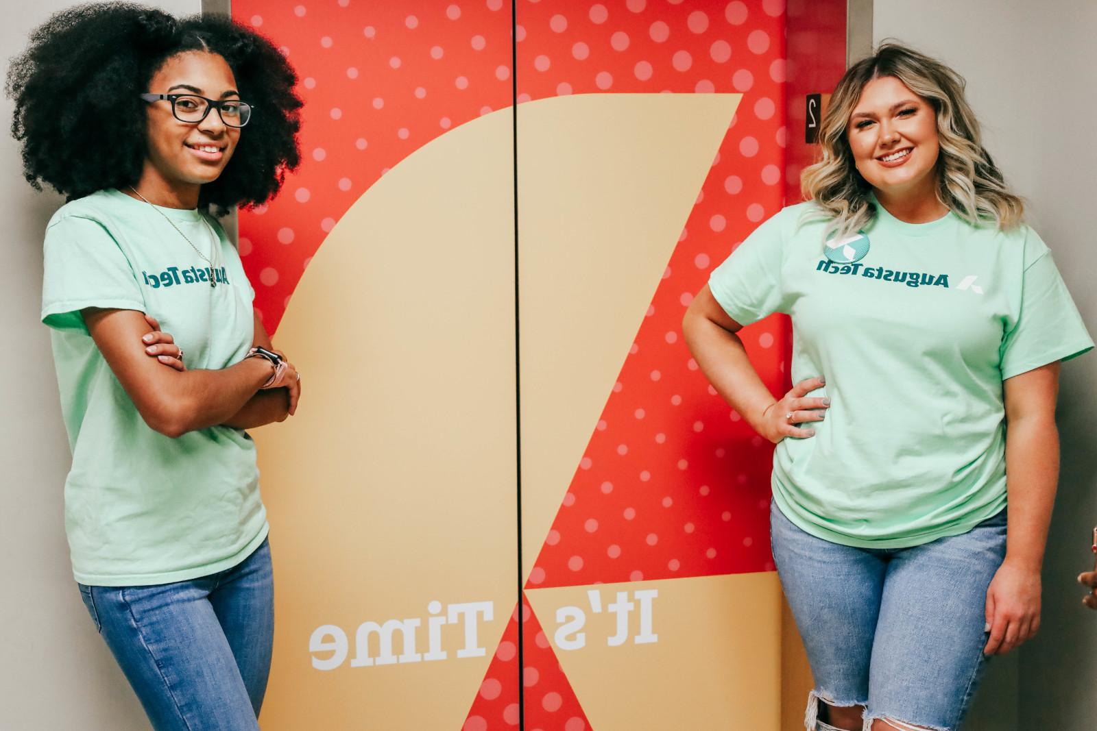 Two female students one a Cacasian female with long blonde hair standing on the left and the other an African American female with shoulder length natural hair on the right, stand wearing mint green Augusta Technical College shirts in front of the 1400 bldg elevator marked by an orange background with a yellow Augusta Technical College A icon and the motto It's Time.